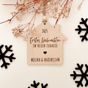 Könnte beinhalten: Ein hölzerner, hausförmiger Weihnachtsschmuck mit der Jahreszahl 2025 oben eingraviert. Der Text "Erstes Weihnachten IM NEUEN ZUHAUSE" und die Namen "MILENA & MAXIMILIAN" sind ebenfalls eingraviert. Der Schmuck hängt an einer Schnur.