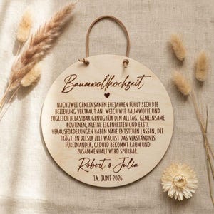 Könnte beinhalten: Runder Holzschild mit dem deutschen Text "Baumwollhochzeit" und einem Herzsymbol. Das Schild enthält gravierten Text mit den Namen "Robert & Julia" und dem Datum "14. JUNI 2026". Eine braune Lederschnur ist zum Aufhängen befestigt.