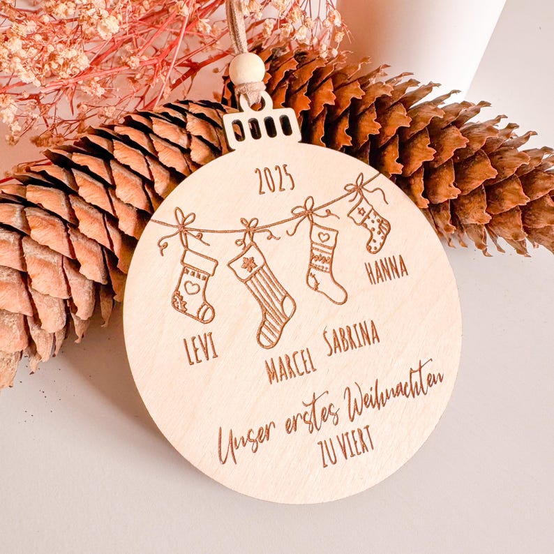 Puede incluir: Adorno navide&ntilde;o redondo de madera clara con calcetines grabados, nombres y el a&ntilde;o 2025. El adorno tiene una cuerda para colgar y presenta los nombres Levi, Marcel, Sabrina y Hanna. Tambi&eacute;n est&aacute; grabado el texto "Unser erstes Weihnachten ZU VIERT".