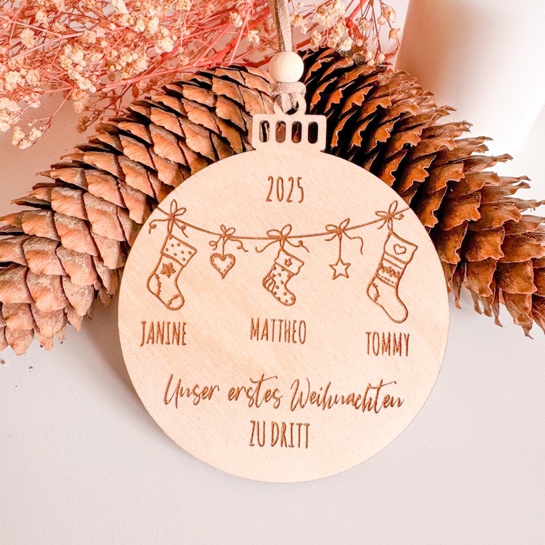 Runde Holz-Weihnachtskugel mit Lasergravur „Unser erstes Weihnachten zu dritt“, Jahr 2025 und drei personalisierten Namen. Motiv mit kleinen Weihnachtsstrümpfen, Herz und Stern. Der Anhänger ist mit veganem Lederband und Holzperle versehen und liegt dekorativ vor Tannenzapfen und getrockneten Blüten.