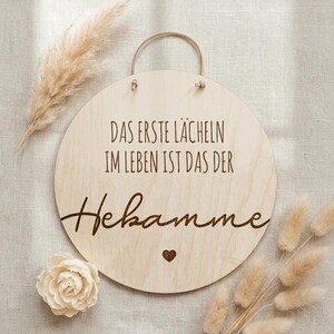 Könnte beinhalten: Ein rundes Schild aus hellem Holz mit der eingravierten deutschen Aufschrift "DAS ERSTE LÄCHELN IM LEBEN IST DAS DER Hebamme" und einem kleinen Herzsymbol unter dem Text. Das Schild verfügt über eine dünne Kordel zum Aufhängen.