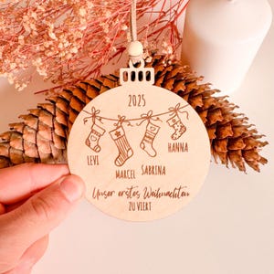 Puede incluir: Adorno navide&ntilde;o redondo de madera clara, grabado con el a&ntilde;o 2025. Presenta cuatro calcetines colgantes con los nombres Levi, Marcel, Sabrina y Hanna. Tambi&eacute;n est&aacute; grabado el texto alem&aacute;n "Unser erstes Weihnachten zu Viert".