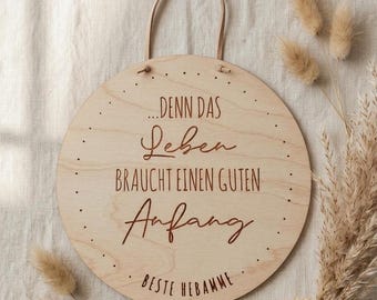 Puede incluir: Un letrero redondo de madera con un asa de cuero marrón claro. El letrero tiene el texto "...DENN DAS Leben BRAUCHT EINEN GUTEN Anfang. BESTE HEBAMME" en una fuente marrón oscuro. Plantas secas rodean el letrero.
