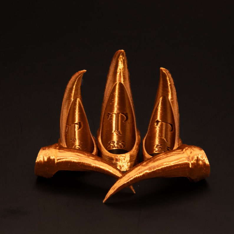 May include: Three gold-coloured claw-shaped finger rings. Each ring has a raised letter "T" on the front.