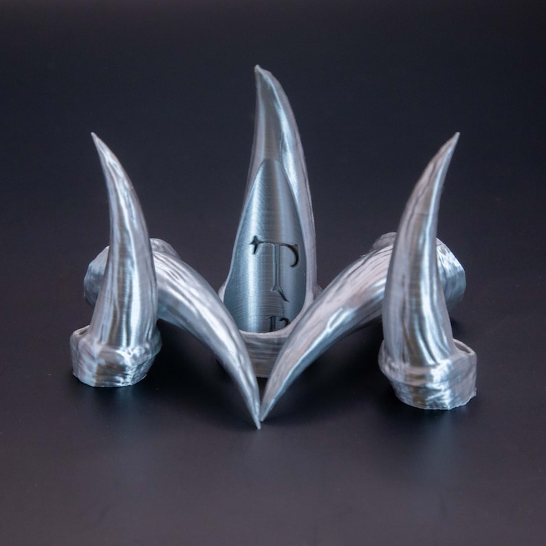 May include: Four silver-coloured, 3D printed, claw-like rings. The centre ring has the letter "T" engraved on it.