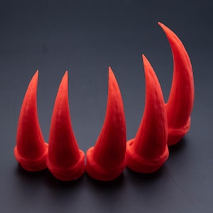 May include: Five bright red plastic horns, resembling claws or fangs, are arranged in a semi-circle on a dark grey surface.