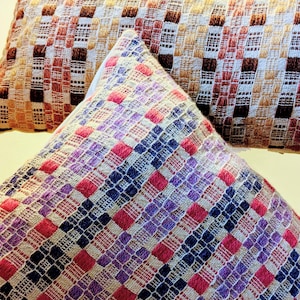 May include: Two decorative cushions with a checkered pattern. The top cushion is brown, white and pink. The bottom cushion is blue, white, pink and purple.