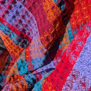 May include: A knitted textile featuring a vibrant, multi-colored pattern with alternating vertical stripes of deep red, bright orange, light purple, and teal. The fabric displays a textured, waffle-like weave with visible yarn loops and varied stitch patterns throughout the material.