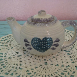 May include: A small, ceramic teapot with a white base and a blue heart design. The teapot has a lid and handle with a speckled purple pattern. It sits on a white, crocheted doily. The teapot is a charming addition to any kitchen.