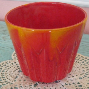 May include: A vibrant, red and orange ceramic planter with a textured, ridged design. The planter has a wide opening and a rounded shape. The exterior features a gradient of colors, transitioning from red to orange. It sits on a white lace doily.