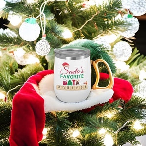 Op de afbeelding: Een witte geïsoleerde wijnbeker met een gouden handvat, met de tekst "Santa's Favorite Data Analyst" in rode, groene en kleurrijke letters. De beker staat tegen een kerstboomachtergrond met ornamenten en lichtjes.