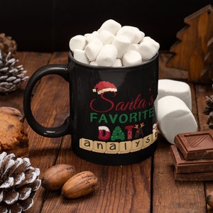 Op de afbeelding: Een zwarte mok gevuld met marshmallows, met de tekst "Santa's Favorite Data Analyst" in een feestelijk ontwerp. De mok is omringd door chocolade, noten en dennenappels op een houten oppervlak, wat een gezellig vakantiethema creëert.