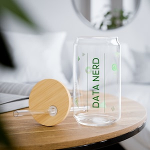Peut inclure: Un gobelet transparent en forme de canette avec les mots "DATA NERD" en vert. Il est doté d'un couvercle en bambou et d'une paille transparente. Le gobelet est posé sur une table en bois. Des icônes graphiques vertes sont dispersées autour du texte.
