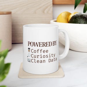 May include: White ceramic mug with the text "POWERED BY Coffee Curiosity Clean Data" in brown. The mug is on a beige coaster. The design includes coffee cup, magnifying glass, and computer icons.
