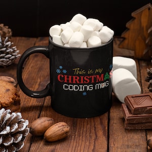 Op de afbeelding: Een zwarte mok gevuld met marshmallows, met de tekst "This is my CHRISTMAS CODING MUG" in rood, wit en groen. De mok staat op een houten oppervlak met dennenappels, noten en chocolade.