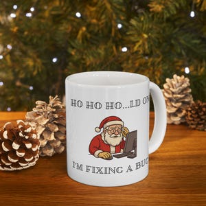 May include: White ceramic mug featuring a cartoon Santa Claus wearing glasses and a red suit, working on a computer. The mug has the text "HO HO HO...LD ON" and "I'M FIXING A BUG".