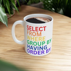 Könnte beinhalten: Weiße Keramik-Tasse gefüllt mit dunklem Kaffee. Die Tasse zeigt den Text "SELECT FROM WHERE GROUP BY HAVING ORDER BY" in Regenbogenfarben. Die Tasse hat einen weißen Henkel und steht auf einer Holzoberfläche.