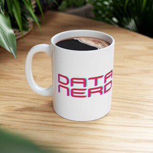 Puede incluir: Taza de cerámica blanca con el texto "DATA NERD" en un diseño en capas de rojo, azul y rosa. La taza está llena de un líquido oscuro, probablemente café o té, y está sobre una superficie de madera.