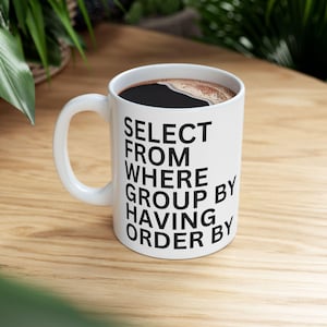 Könnte beinhalten: Weiße Keramik-Kaffeetasse gefüllt mit dunkler Flüssigkeit, wahrscheinlich Kaffee. Die Tasse hat schwarzen Text: "SELECT FROM WHERE GROUP BY HAVING ORDER BY". Die Tasse steht auf einer hellen Holzoberfläche.