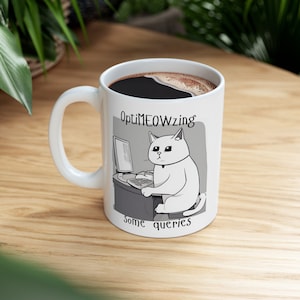 May include: White ceramic coffee mug featuring a black cat illustration. The cat is sat at a computer and the text "OptiMEOWzing some queries" is printed on the mug.