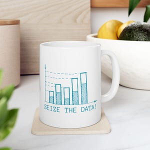Puede incluir: Taza de cerámica blanca con un diseño de gráfico de barras turquesa y el texto "SEIZE THE DATA!". La taza tiene un asa curva y está sobre un posavasos beige. El diseño sugiere un tema de datos o negocios.