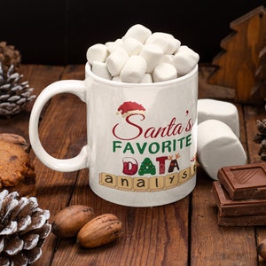 Op de afbeelding: Een witte keramische mok gevuld met marshmallows, met de tekst "Santa's Favorite DATA analyst" in feestelijke kleuren. De mok staat op een houten oppervlak met chocoladeblokjes, dennenappels en pecannoten.