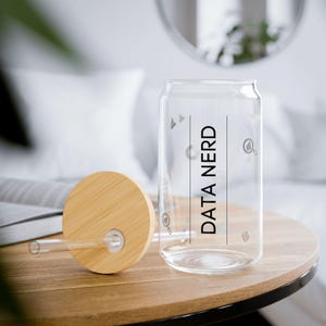 Puede incluir: Un vaso transparente en forma de lata con las palabras "DATA NERD" impresas verticalmente en negro. Tiene una tapa de bambú y una pajita transparente. El vaso está sobre una mesa de madera.