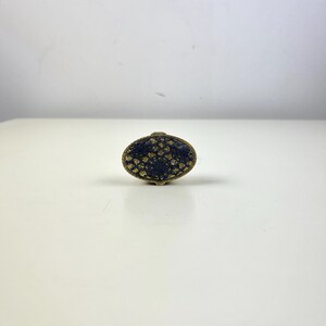 May include: An oval-shaped cabinet knob with a dark blue surface and a gold-colored, patterned design. The knob has a small, gold-colored base and a matching top. The hardware is likely for furniture or decorative use.