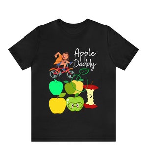 Puede incluir: Camiseta negra con una ilustración de dibujos animados de una persona montando en bicicleta con el texto "Apple Daddy" y un grupo de manzanas, incluida una manzana verde con gafas.