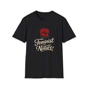 Puede incluir: Camiseta negra con un diseño de rosas rojas y el texto "Feminist by Nature" en cursiva. El gráfico está centrado en la parte delantera de la camiseta de manga corta. La camiseta está hecha de tela suave.