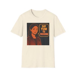 Può includere: T-shirt color crema con grafica di una donna in blazer e la scritta "SAY YUM TO FREEDOM" in un fumetto. Il design ha uno stile retrò con colori arancione e nero, che ricorda un fumetto.