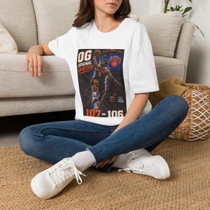 May include: A white short-sleeved t-shirt featuring a graphic print of a basketball player in a blue and orange jersey performing a slam dunk. The text reads OG ORIGINAL GAMESTER, NEW YORK KNICKS, and the score 107-106 in bold orange and white.