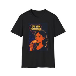 Può includere: T-shirt nero con una grafica di una donna che tiene in mano uno snack con la scritta "YUM". La grafica include anche il testo "SAY YUM TO FREEDOM" in un fumetto. La pelle della donna è arancione e lo sfondo è blu scuro.