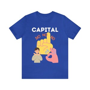 Puede incluir: Una camiseta azul real con el texto "CAPITAL" en letras blancas. Debajo del texto hay una mano amarilla con un dedo apuntando hacia arriba y las palabras "NO NO NO" escritas a su alrededor. Una persona de dibujos animados con cabello castaño se muestra con los brazos cruzados, y una mano rosa con una señal de OK se muestra a la derecha de la mano amarilla.