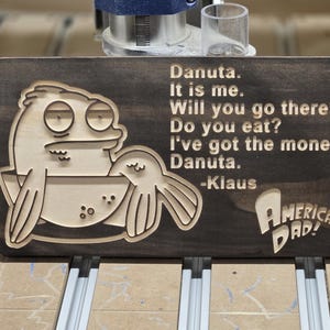 Peut inclure: Plaque en bois avec un personnage de dessin animé et du texte de la série animée "American Dad!". La plaque présente le personnage Klaus et la citation "Danuta. C'est moi. Iras-tu là-bas ? Manges-tu ? J'ai l'argent. Danuta."