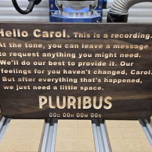 May include: A dark brown wooden sign with engraved text that reads "Hello Carol. This is a recording." and other text. The sign also has the word "PLURIBUS" engraved on it. The sign is on a metal surface.