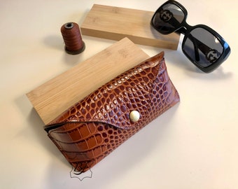 May include: Brown leather sunglasses case with a crocodile skin texture, secured with a white button. Black sunglasses with a square frame and the Gucci logo are also visible. A spool of thread and wooden blocks are in the background.