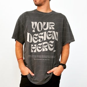 May include: A grey t-shirt with a vintage-style graphic that says "YOUR DESIGN HERE". The text is in white and has a retro font. The shirt is being modelled by a person with a beard and a watch on their wrist.