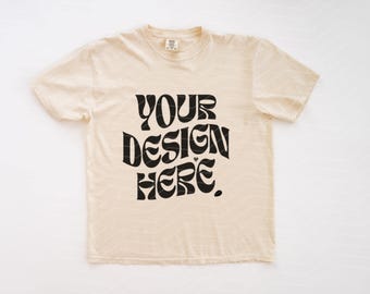 May include: A short-sleeved, crew-neck t-shirt in a light beige color. The center of the shirt features bold, black, wavy-style lettering that reads "YOUR DESIGN HERE." The fabric has a subtle, light-colored chevron pattern throughout the material.
