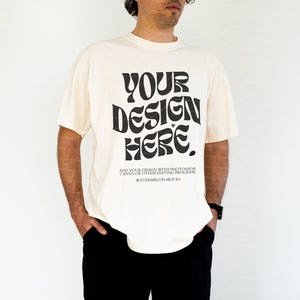 Könnte beinhalten: Ein cremefarbenes Kurzarm-T-Shirt mit dem schwarzen Text "YOUR DESIGN HERE." und kleinerem Text darunter, der "ADD YOUR DESIGN WITH PHOTOSHOP, CANVA OR OTHER EDITING PROGRAM" und "WATERMELON MOCKS" auf der Vorderseite lautet.