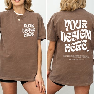 以下が含まれることがあります： 「YOUR DESIGN HERE」と白で書かれた茶色のTシャツ。レトロなフォントスタイルのグラフィックです。Tシャツはモデル着用です。