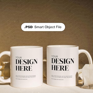 May include: Two white ceramic mugs with black text that reads "Your Design Here". The mugs are on a white surface with a tea bag in the foreground.