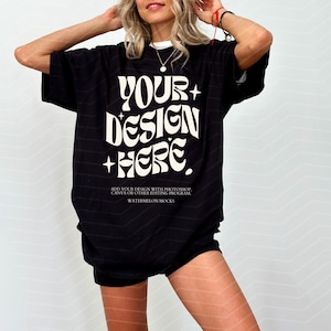 Pode incluir: Uma t-shirt preta oversize com o texto branco "YOUR DESIGN HERE" numa fonte estilizada e pequenos detalhes de estrelas. Abaixo, um texto branco mais pequeno diz "ADD YOUR DESIGN WITH PHOTOSHOP, CANVA OR OTHER EDITING PROGRAM. WATERMELON MOCKS."