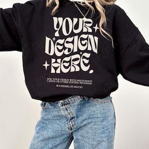 May include: A black sweatshirt with the text "YOUR+DESIGN+HERE" in white. The text is in a retro font style. The sweatshirt is being modelled by a person wearing blue jeans.
