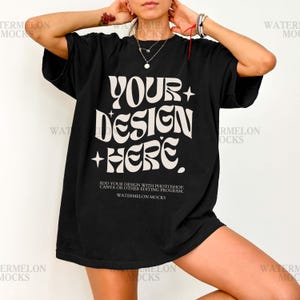 Op de afbeelding: Zwart oversized T-shirt met de tekst "YOUR DESIGN HERE" in een witte, retro stijl. Het shirt bevat ook de tekst "ADD YOUR DESIGN WITH PHOTOSHOP, CANVA OR OTHER EDITING PROGRAM. WATERMELON MOCKS".