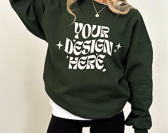 May include: A dark forest green long-sleeve crewneck sweatshirt featuring the white text "YOUR DESIGN HERE" in a wavy, stylized font, accented by four-pointed star symbols on the left and right sides of the central text.