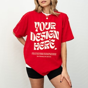 May include: Red oversized t-shirt with short sleeves featuring the text "YOUR DESIGN HERE" in white. The text is in a retro, psychedelic font. The text below the design reads "ADD YOUR DESIGN WITH PHOTOSHOP, CANVA OR OTHER EDITING PROGRAM. WATERMELON MOCKS."