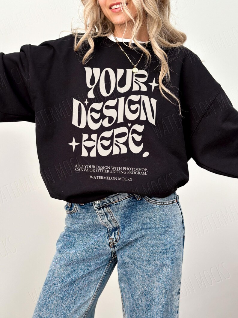 May include: A black sweatshirt with the text "YOUR+DESIGN+HERE" in white. The text is in a retro font style. The sweatshirt is being modeled by a person wearing blue jeans.