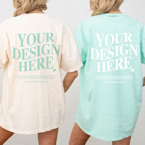 Op de afbeelding: Twee oversized T-shirts, een ivoor en een lichtblauw, met de tekst "YOUR DESIGN HERE" gedrukt op de achterkant. De tekst is lichtgroen.
