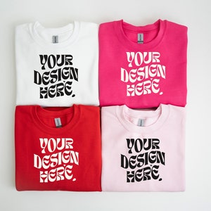 May include: Four folded sweatshirts in different colors: white, pink, red, and light pink. Each sweatshirt has the text "YOUR DESIGN HERE." printed in black.